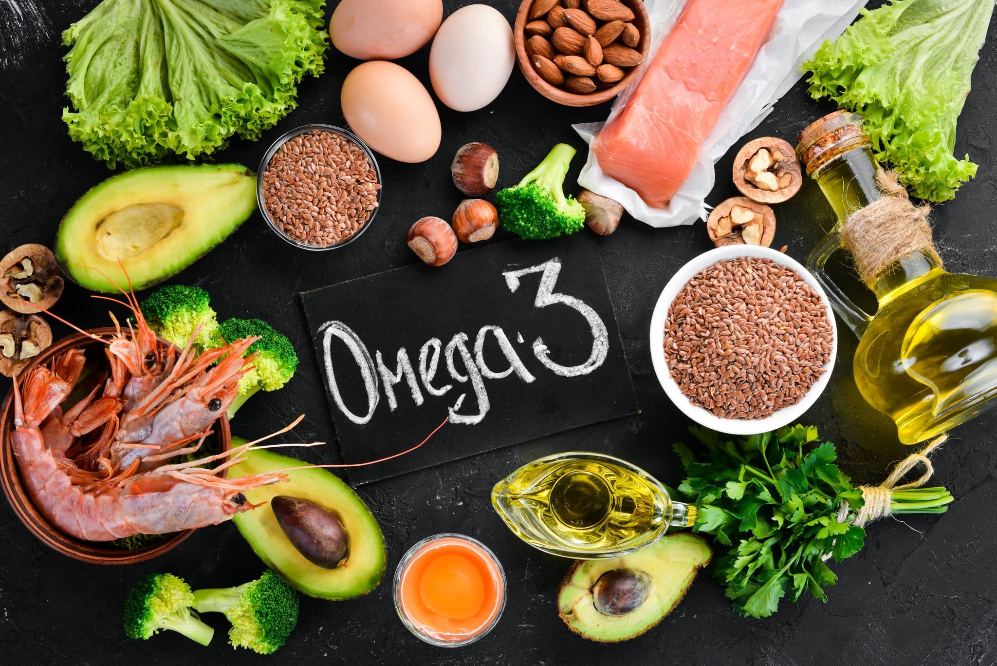 "Omega 3" steht auf einer schwarzen Tafel, darum herum Nahrungsmittel, die viel Omega 3 enthalten, wie Nüsse, Avocado, Lachs, Eier oder Öl.