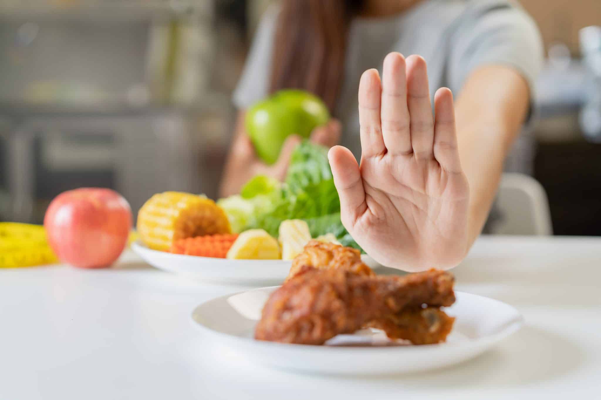 Im Vordergrund ein Teller mit Hühnchen, den eine Hand symbolische wegdrückt, im Hintergrund ein Teller mit Obst und Gemüse, vor einem verschwommenen Oberkörper.