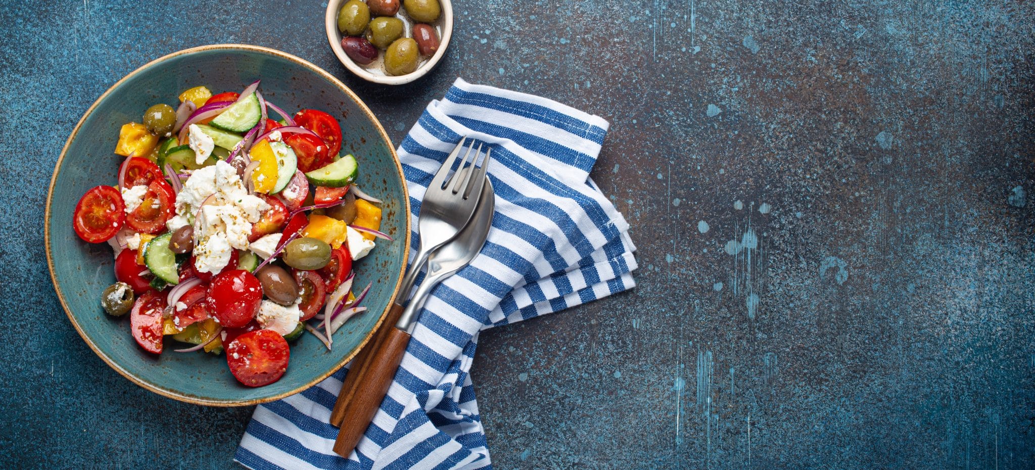 Teller mit griechischem Salat mit Feta-Köse und buntem Gemüse, daneben ein kleiner Teller Oliven und eine blau-weiß-gestreifte Serviette, auf der eine Gabel und ein Löffel liegen.