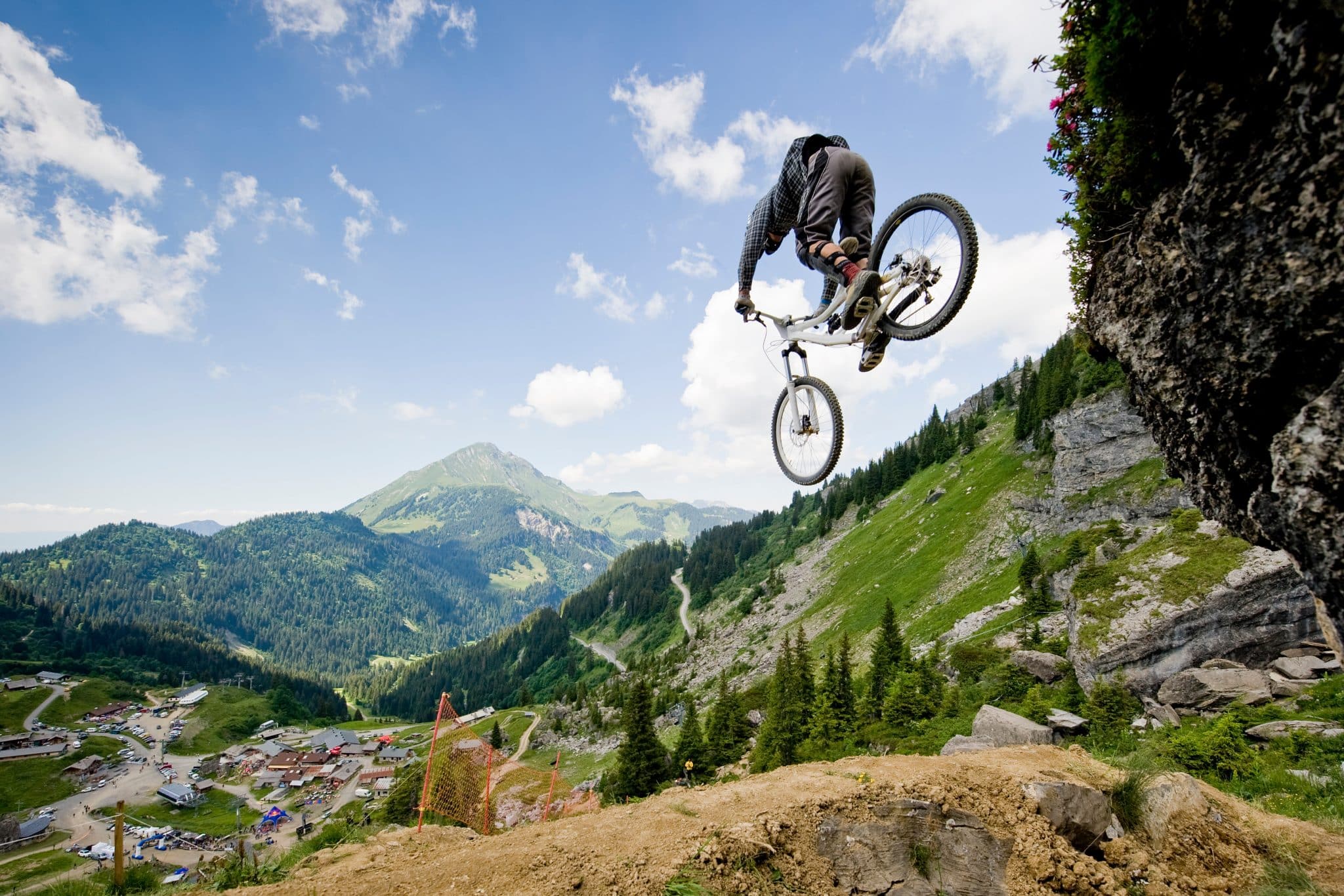 Mountainbiker springt vor Alpenlandschaft von einem Felsen; im Hintergrund sieht man Zelte einer Sportveranstaltung.