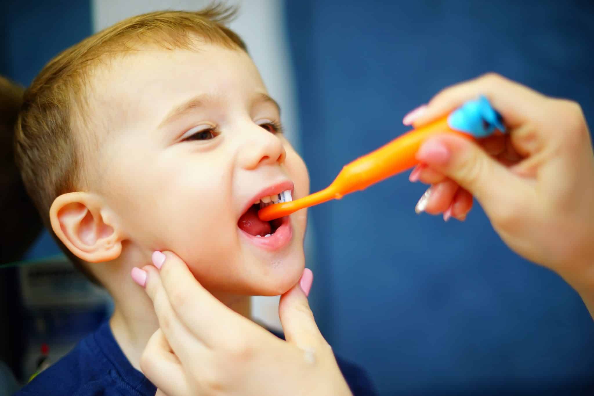 Wir sehen den Kopf eines Kinds mit offenem Mund; dazu Frauenhände, die eine Zahnbürste halten und ihm die Zähne putzen.
