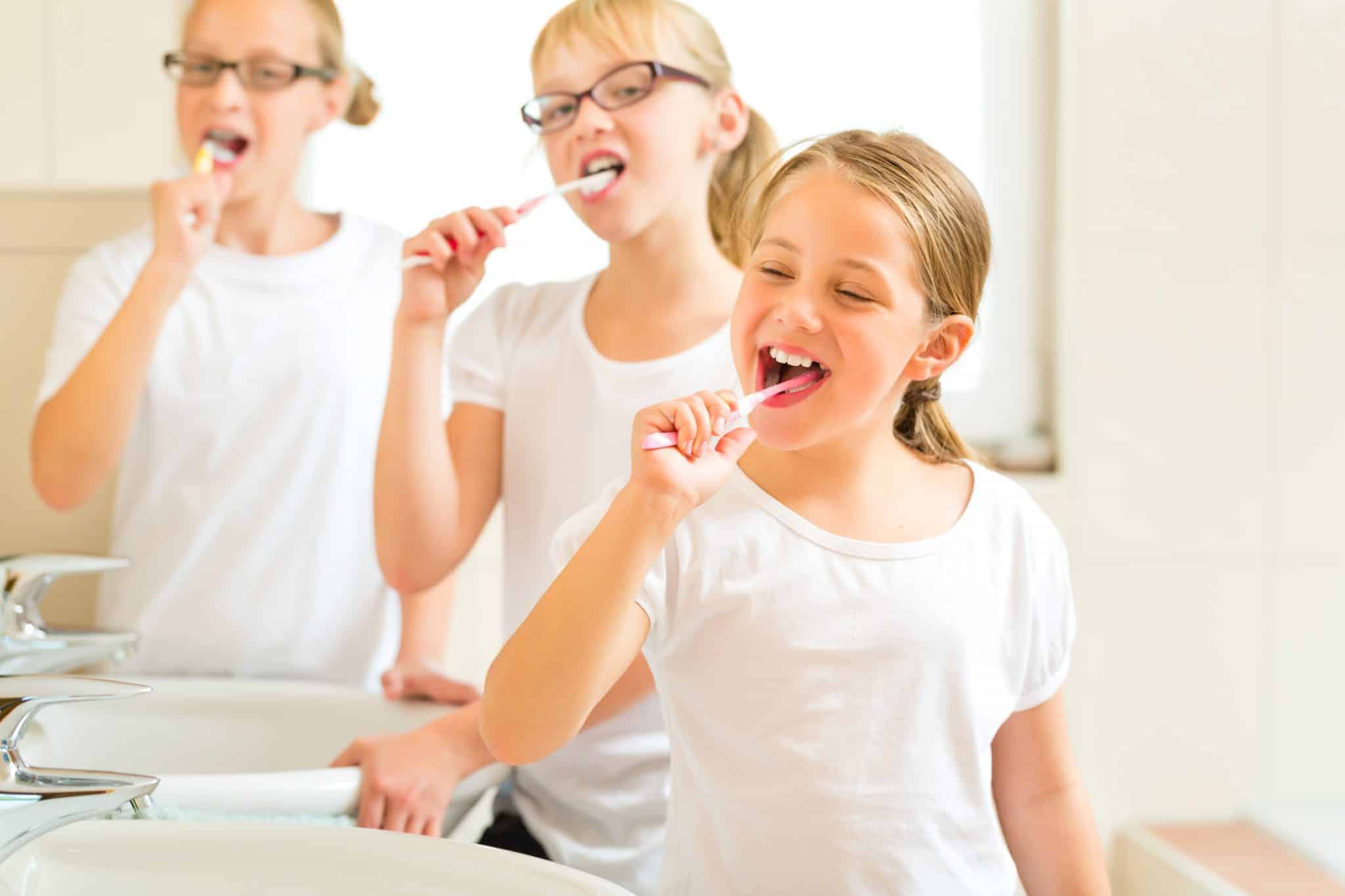 3 Mädchen in weißen T-Shirts stehen in einem Bad mit zwei Waschbecken und putzen sich die Zähne