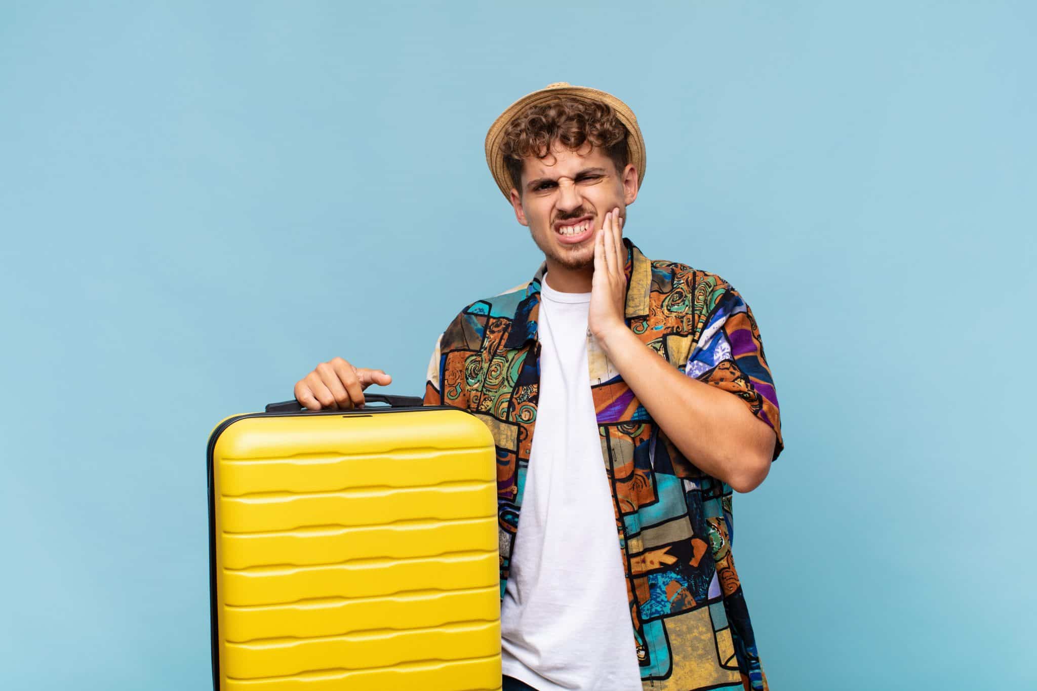 Junger Mann im Urlaubs-Outfit und mit Koffer hält sich die Backe vor Zahnschmerzen