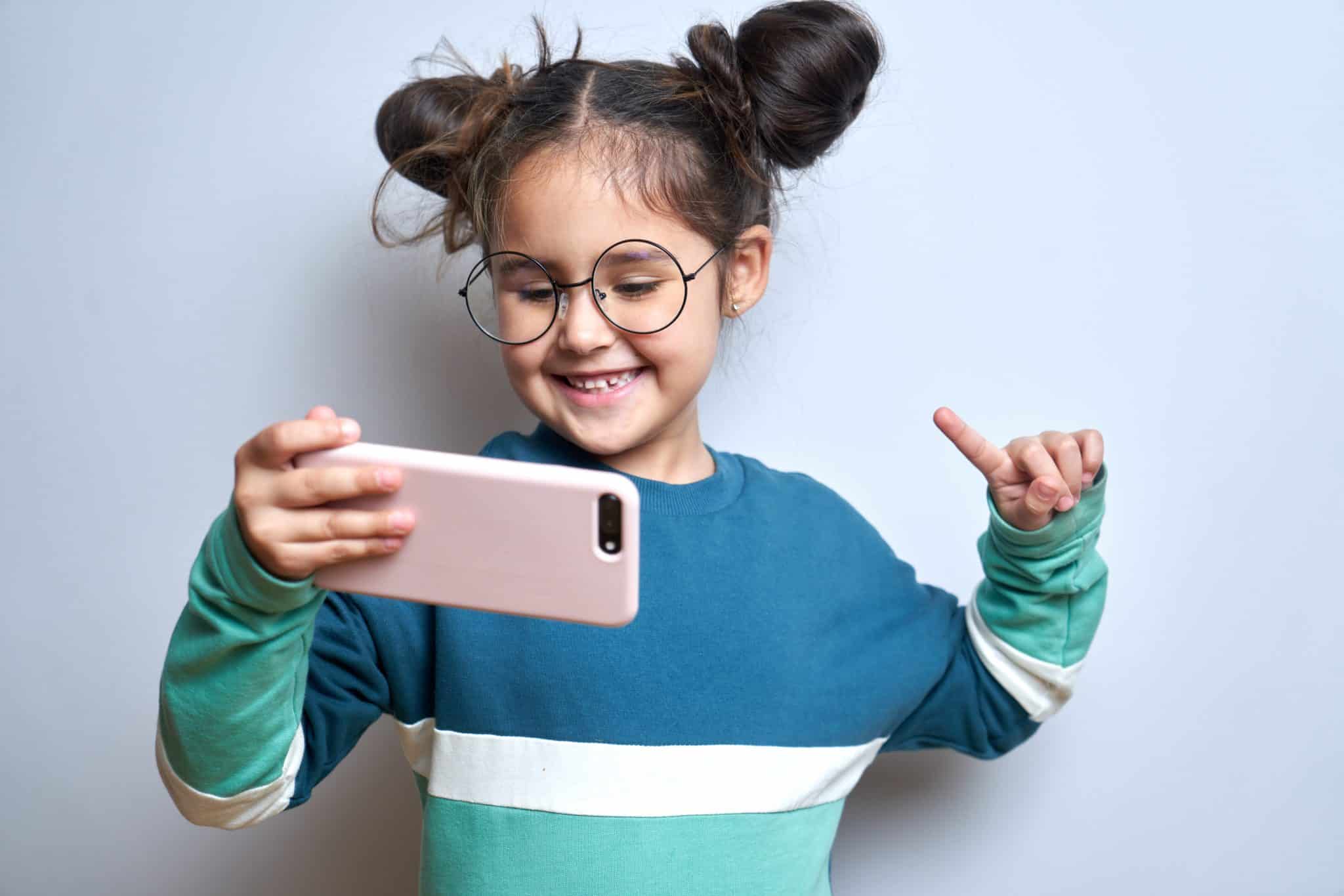 Ein lachendes Mädchen mit großer Brille und einem Handy in der Hand vor blauem Hintergrund.