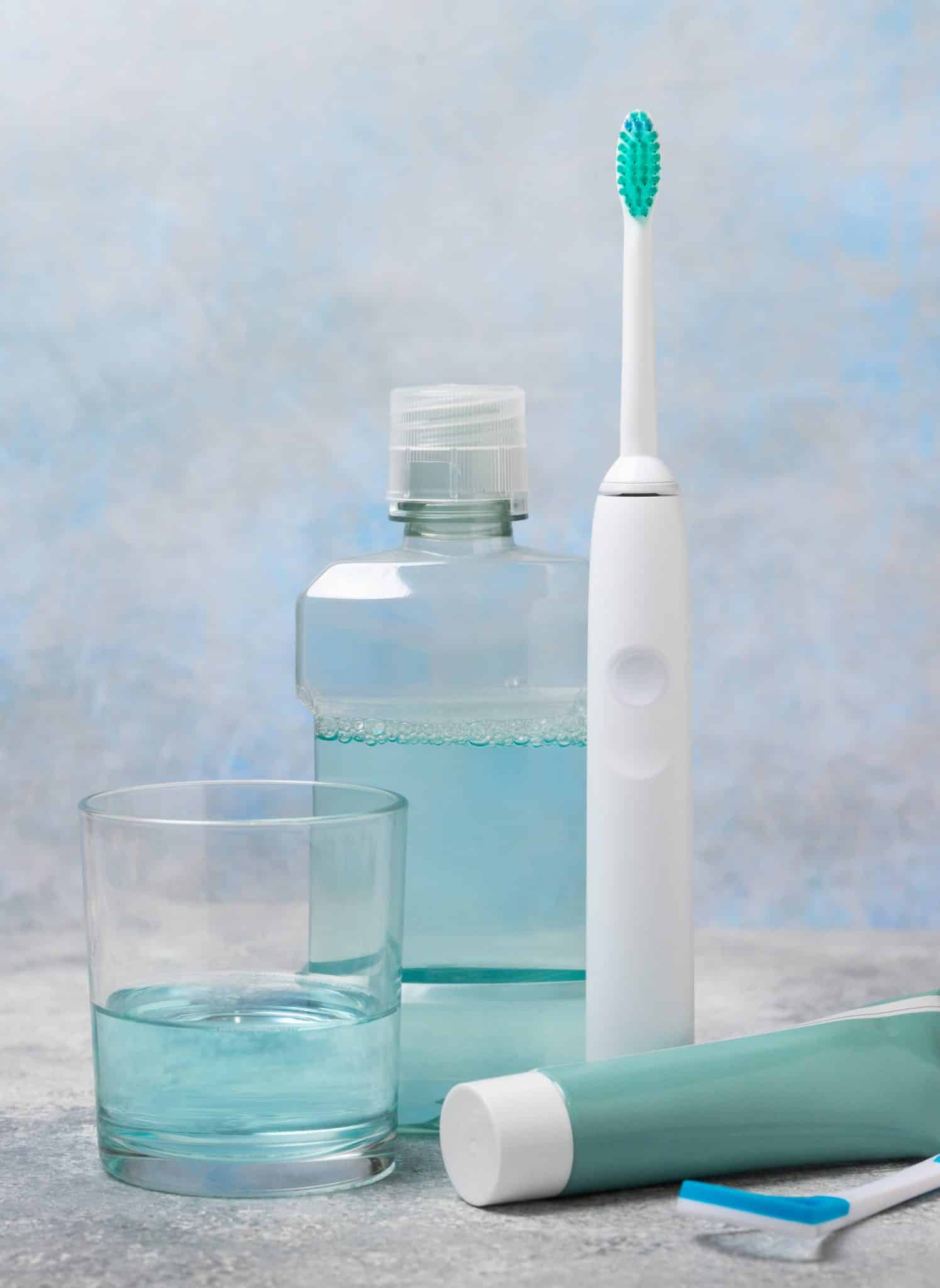Zahnputz-Ausrüstung auf Fliesenablage: Wasserglas, Mundspülung, Ultraschallzahnbürste, Zahnpasta und Zungenschaber