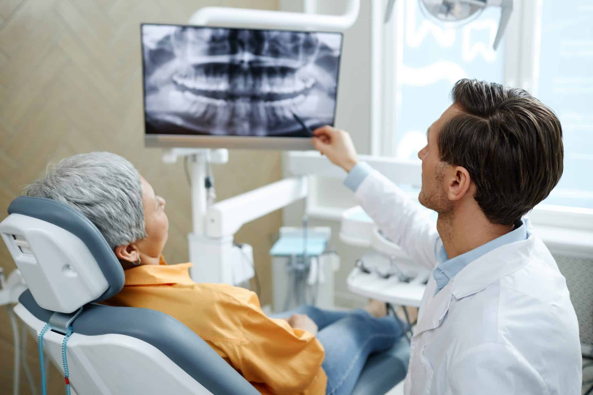 Ein Zahnarzt erklärt einer älteren Frau auf dem Zahnarzt-Behandlungsstuhl ihr Röntgenbild, das auf einem Bildschirm im Praxisraum angezeigt wird.