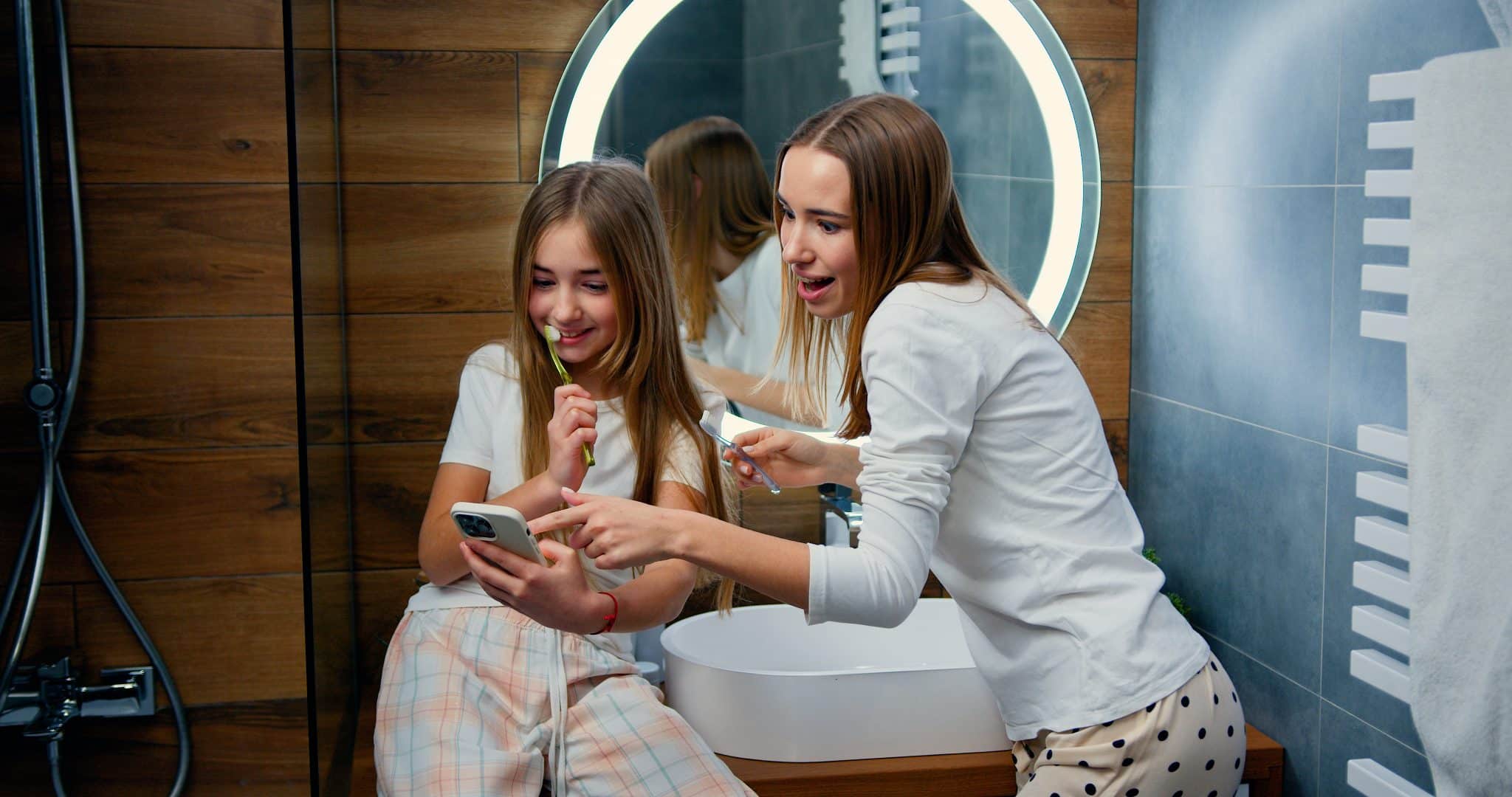 Zwei blonde Mädchen schauen beim Zähneputzen aufs Handy.
