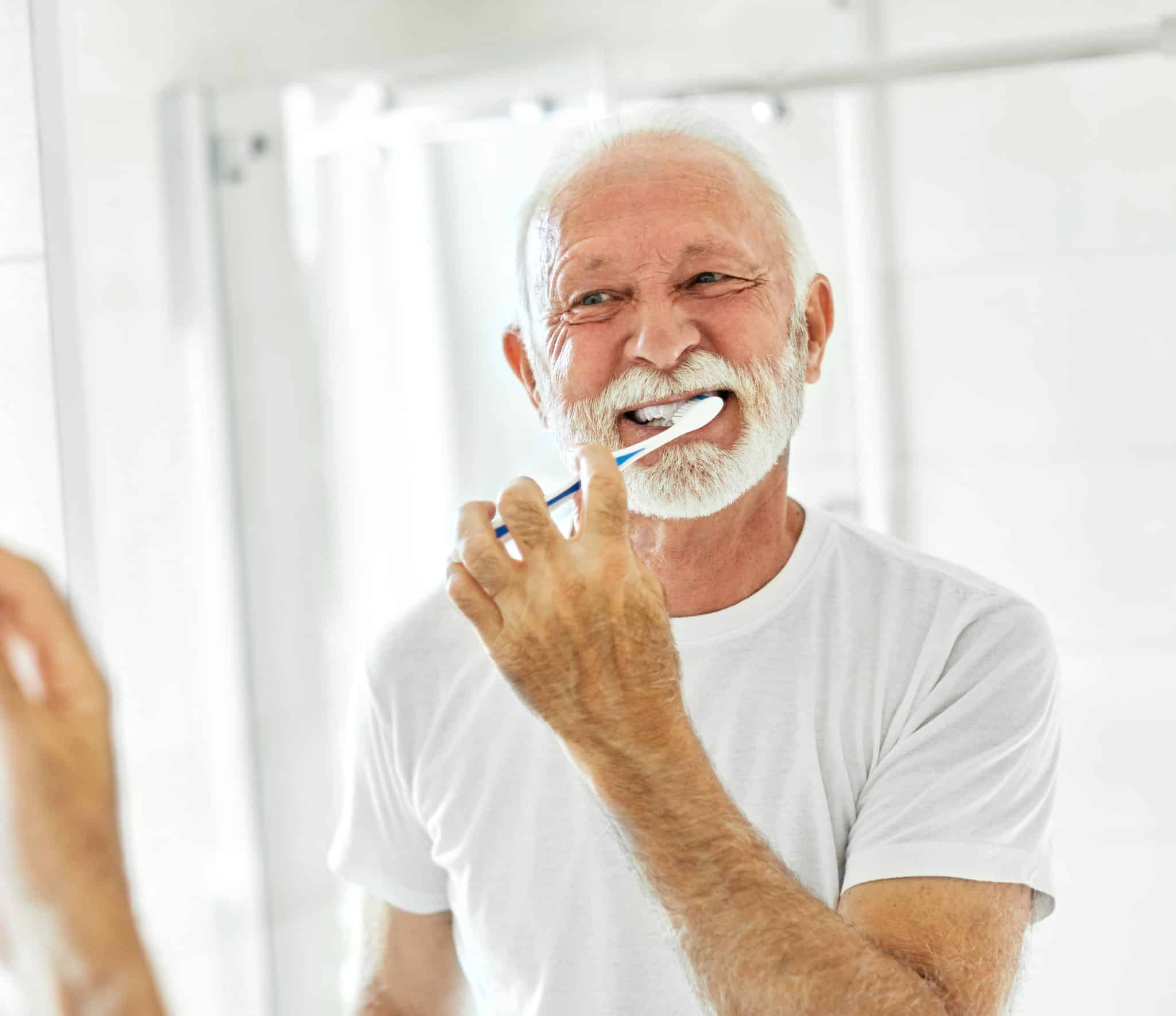 Ein älterer Mann putzt sich die Zähne im Badezimmer vor dem Spiegel.