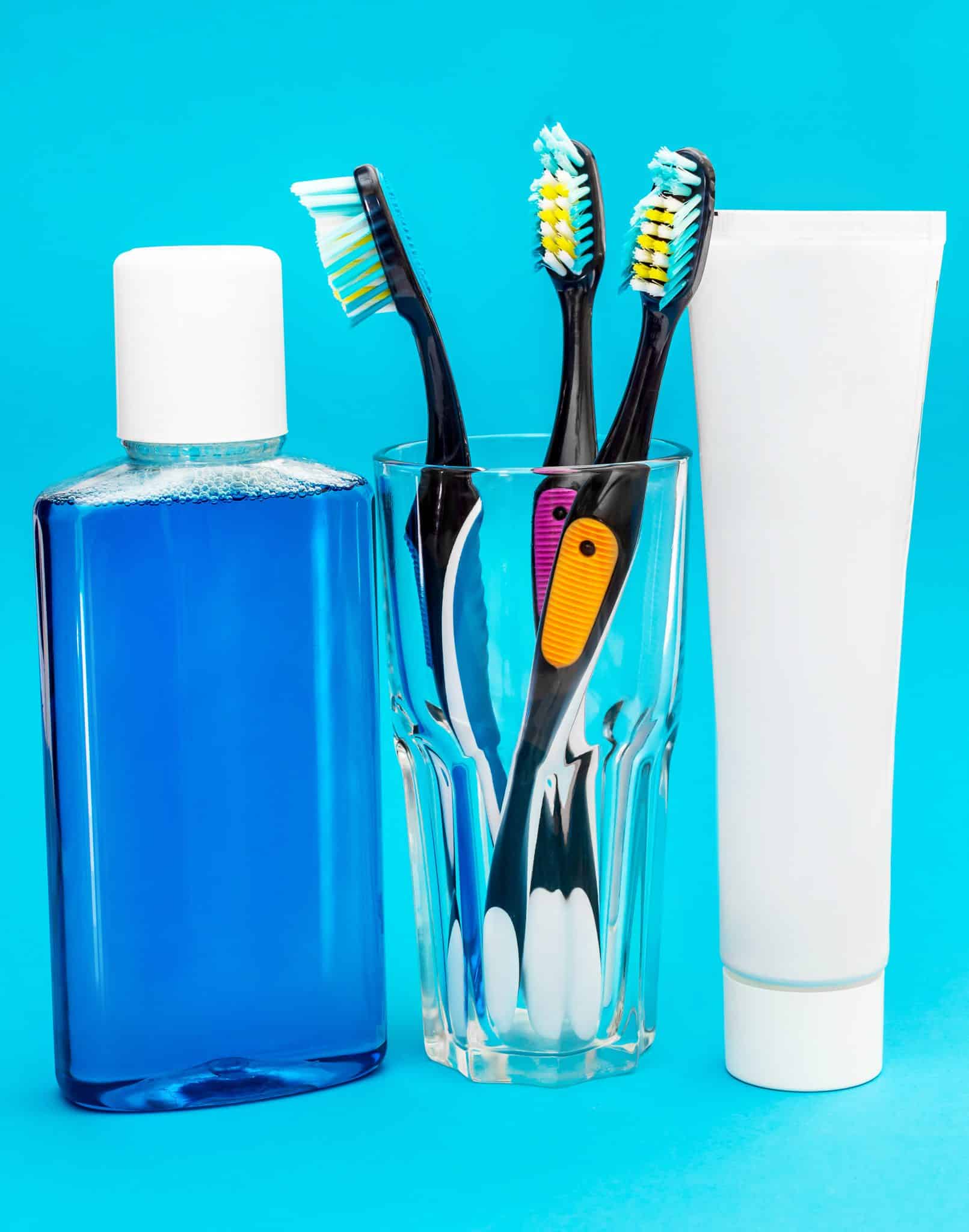 Das Foto zeigt ein Zahnpflegeset mit Zahnbürsten in einem Glas, Zahnpasta und Mundspülung auf hellblauem Hintergrund.