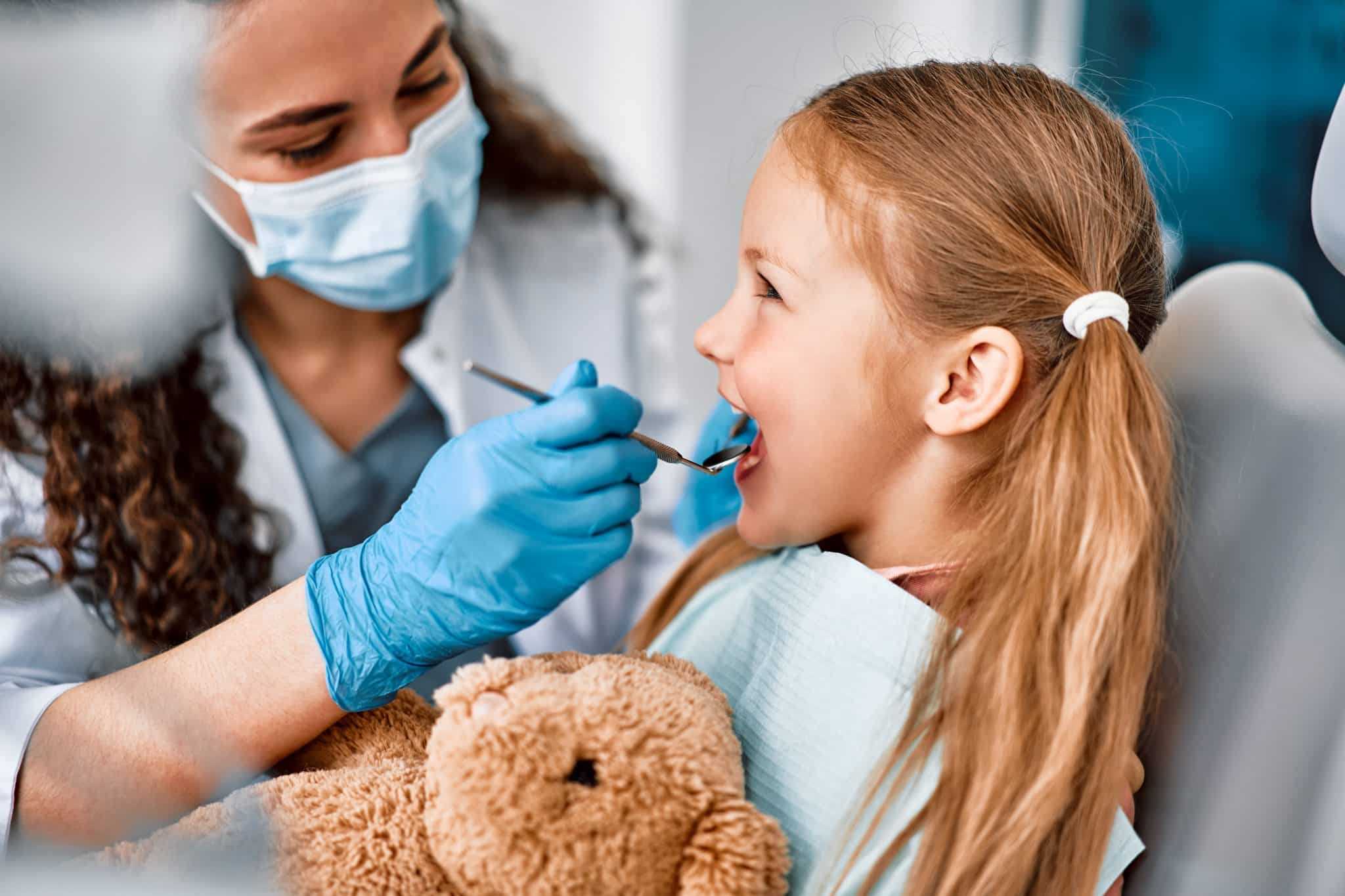 Das Foto zeigt eine Zahnärztin bei der Untersuchung der Zähne eines kleinen Mädchens mit seinem Teddy.