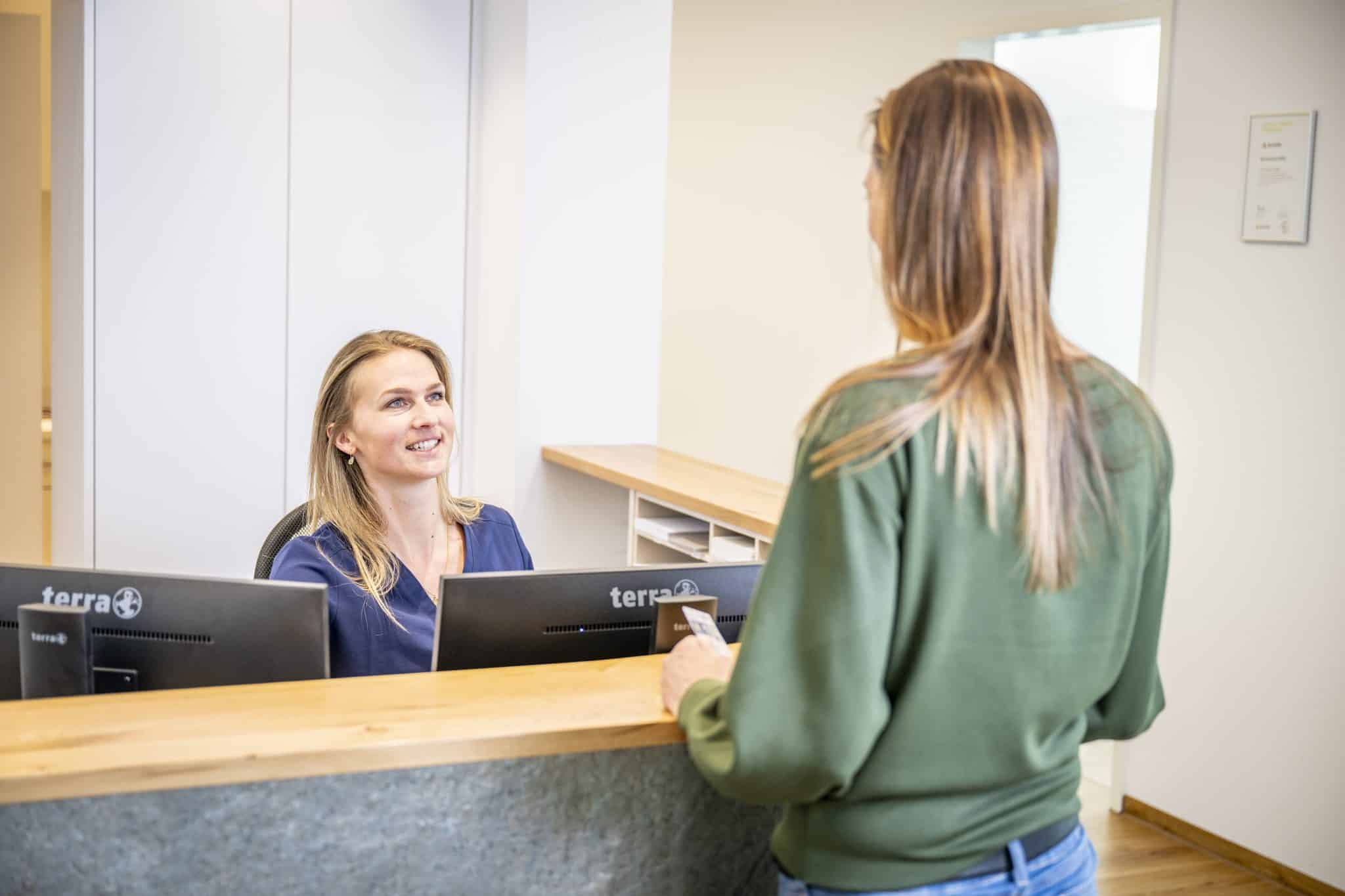 Eine Patientin mit ihrer Gesundheitskarte im Gespräch mit der freundlichen und aufmerksamen Mitarbeiterin am Empfang der Zahnarztpraxis.