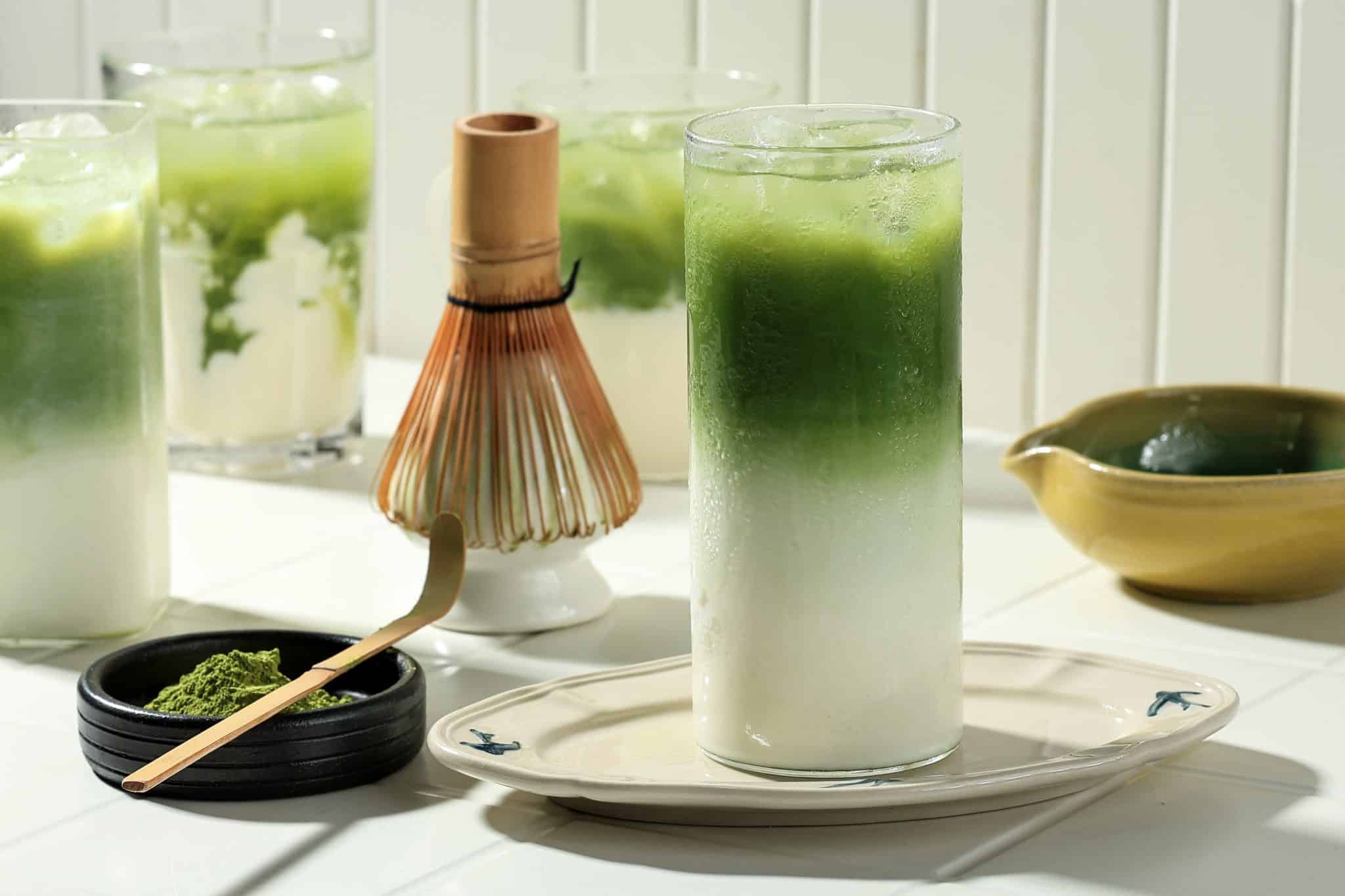 Das Foto zeigt ein Glas eisgekühlten grünen Matcha-Tee mit Milch, Schale mit grünem Matcha-Pulver und Matcha-Löffel.