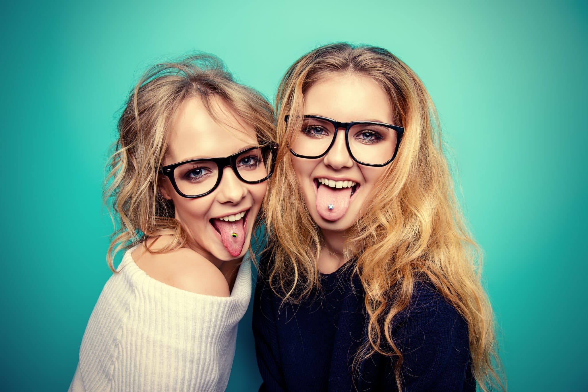 Zu sehen sind zwei junge Frauen mit langen, blonden Haaren und großen Brillen die ihr Zungenpiercing mit herausgestreckter Zunge zeigen.